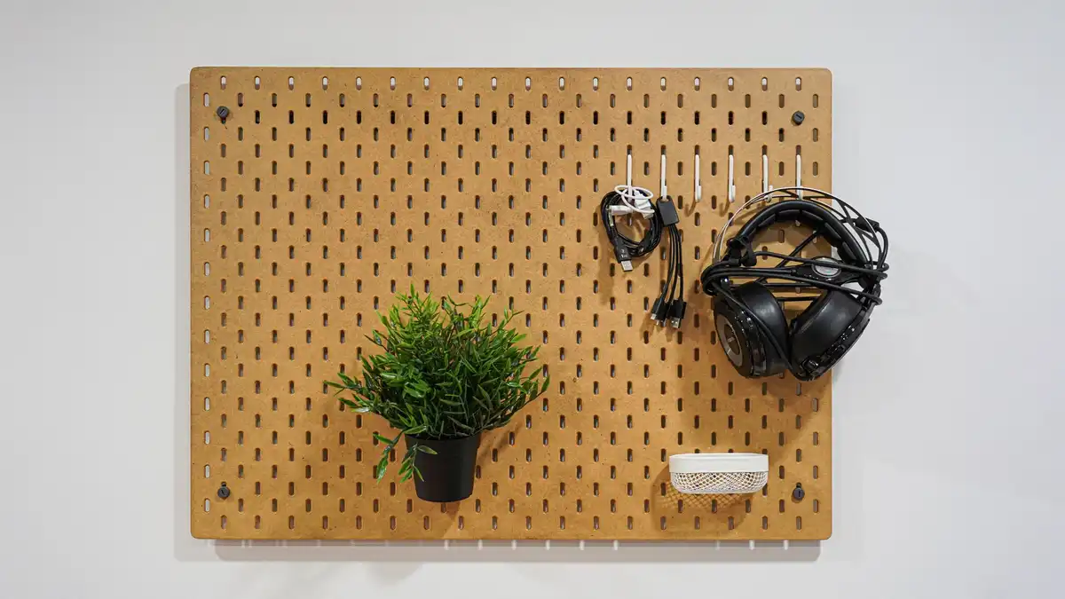 A wooden pegboard on a white wall holding a small plant, headphones and cables