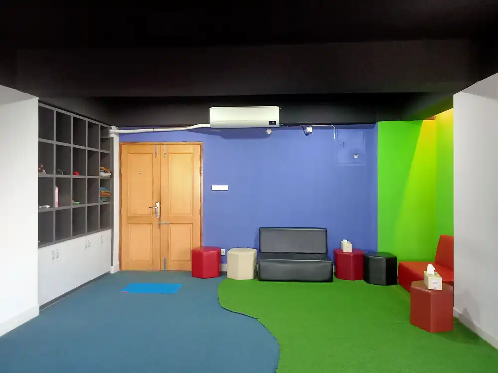 A colourful office corner with a blue wall, green floor, cube shelves and small red stools