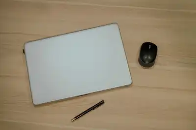 A closed white laptop, a wireless mouse and a pen on a light wooden desk, seen from above