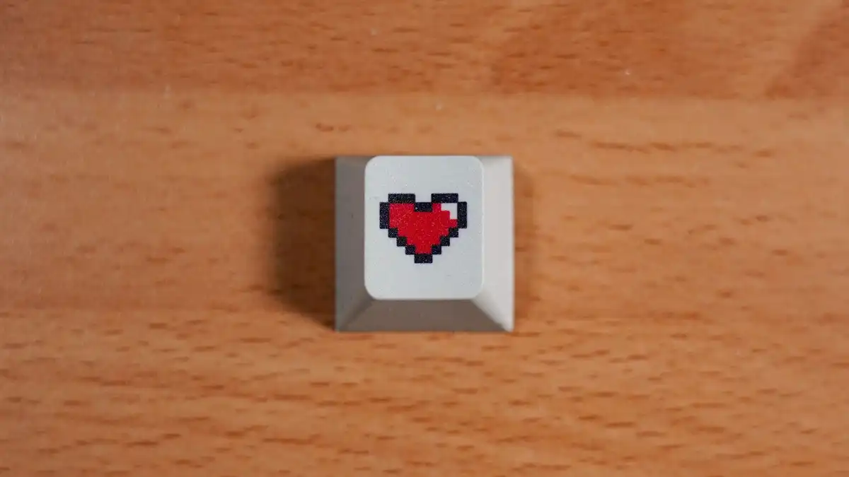 A single grey keycap printed with a red pixel heart on a wooden table