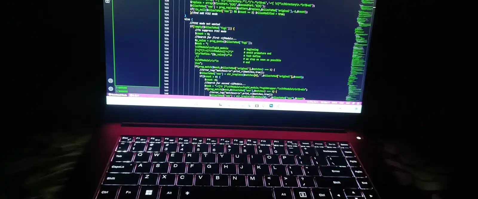 A laptop in a dark room, its screen showing green code and its keyboard lit