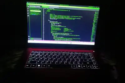 A laptop in a dark room, its screen showing green code and its keyboard lit